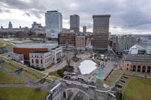 Aerial view over Quebec City, with the stone rampart and the Porte Saint-Jean gate running across the foreground, an outdoor skating rink below, and downtown office towers behind.