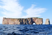 The Rocher Perce seen from the water, a long limestone cliff rising from the sea with an arch pierced through one end and a separate rock pillar standing beyond it.