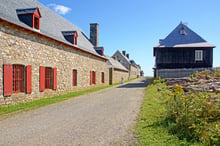 A gravel lane between stone buildings with red shutters and slate roofs, a woman in eighteenth-century dress walking away, blue sky