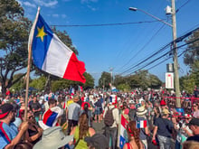 A crowd filling a street under a large blue, white and red Acadian flag with a yellow star, on a sunny day