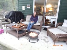 A woman sitting on a patio sofa beside a lit fire bowl, a tray of honey jars on the low table in front of her and snow at the edge of the concrete.