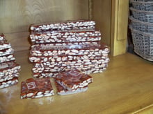 Stacks of cling-wrapped dark brown nougat bars studded with whole almonds on a wooden shop shelf.