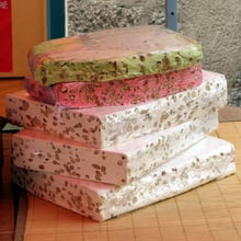 Cling-wrapped slabs of white almond nougat topped by a pistachio-green slab and a pink slab, on a bamboo mat.