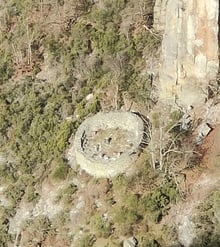 A circular stone-walled enclosure on a wooded slope beside a rock face.