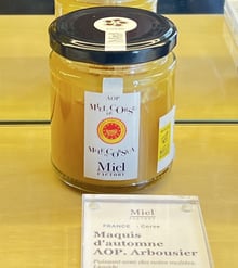 A jar of pale golden honey with an AOP Miel de Corse label, above a shop card reading Maquis d'automne AOP. Arbousier.