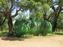 A large green cylindrical water tank under spreading cork oaks beside a dirt clearing.