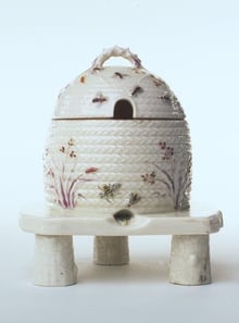 A white porcelain honey pot shaped as a straw beehive, with a domed lid and an entrance hole, painted with grasses and bees, standing on a small stand with log-shaped legs.