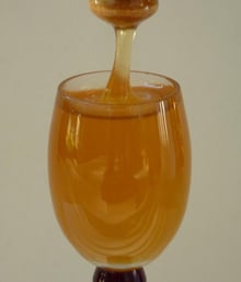 A stemmed glass filled with liquid honey, a wooden dipper above it trailing a thread of honey into the glass.