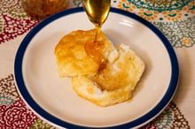 Honey running from a spoon onto a split biscuit on a white plate.