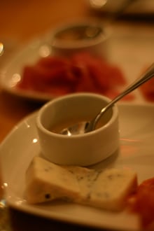 A wedge of blue-veined cheese on a white plate beside a small dish of honey with a spoon.