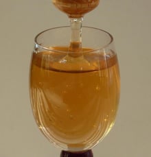 A stemmed glass of clear pale amber honey with a wooden dipper standing in it.