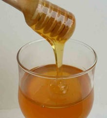 Clear golden honey running off a wooden honey dipper into a stemmed glass half full of honey.
