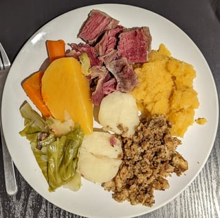 White plate holding boiled salt beef, cabbage, carrots, turnip, potatoes and bread stuffing.