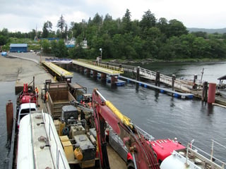 Heavy trucks and an excavator loaded on a vessel deck beside a floating dock and forested shore.