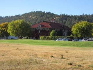 A large red-roofed brick building with ivy on its walls, seen across mown grass with wooded hills behind.