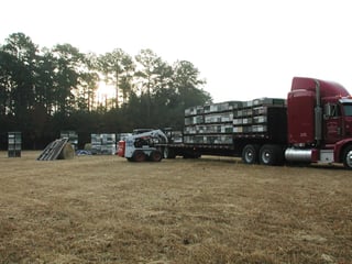 A skid-steer loader lifting pallets of stacked beehives onto a red flatbed semi-trailer in a dry field at dusk, more pallets waiting on the grass.