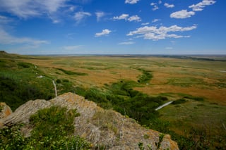 A lichen-covered sandstone ledge in the foreground, a trail and boardwalk on the slope below, and rolling green and gold grassland stretching to a flat horizon under a blue sky with scattered cloud.