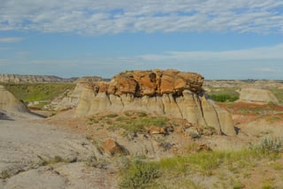 Pale grey and tan striped badland slopes topped by a rust-brown rock cap, red-brown flats and green sage in the foreground, more striped hills and a green valley beyond under a blue sky with broken cloud.