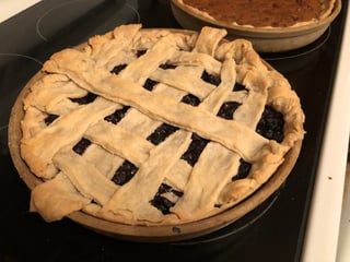 A whole lattice-crust pie in a round stoneware dish on a black stovetop, dark purple berry filling showing between the pastry strips, a second pie behind it.