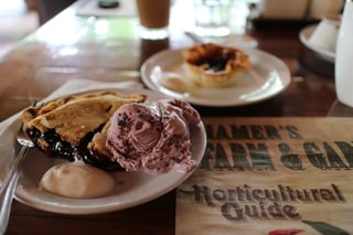 A wedge of dark purple berry pie with a scoop of pink berry ice cream and a spoonful of cream on a white plate, a small butter tart on a plate behind, on a wooden cafe table.