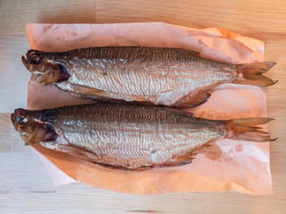 Two whole smoked fish with gold-bronze skin laid head to tail on pink butcher paper on a pale wooden table, photographed from above.