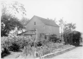 A two-storey clapboard house with a single-storey extension, an early motor truck parked at the door, and a vegetable garden in front.
