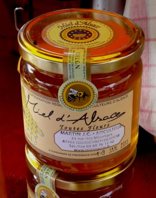 A single jar of honey labelled Miel d'Alsace toutes fleurs with a round IGP sticker on its lid, standing on a red cloth.