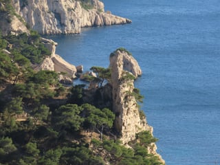 A narrow limestone inlet with clear turquoise water between steep white cliffs covered in low pine and scrub.