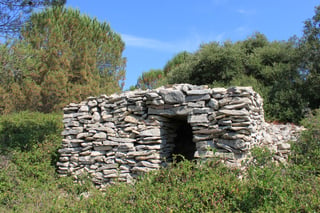 A small corbelled drystone hut with a domed roof standing in low Mediterranean scrub, built without mortar from flat limestone slabs.
