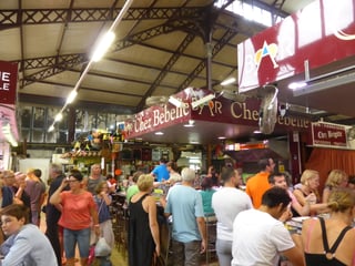 The interior of an iron and glass market hall, riveted roof trusses overhead, with a long dark red sign reading Bar Chez Bebelle above a grill counter at the centre.