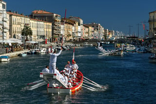 A jouster in white standing alone on a beam projecting from the stern of a red barque, shield raised and lance levelled, over a canal lined with spectators.
