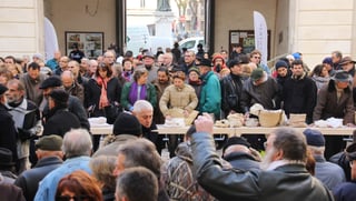 A winter crowd in coats and caps pressed against a long bare trestle under an arcade, straw panniers and closed white cloth sacks on the table.
