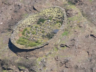 Seen from above, an oval dry-stone enclosure on a bare hillside with rows of small hives inside.
