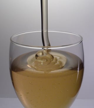 Pale amber honey falling from a dipper and coiling into a clear stemmed glass half full of honey.