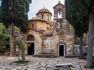 A small stone-and-brick Byzantine church with a tiled dome and a bell-cote, framed by tall cypresses.
