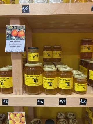 Jars with yellow labels reading Miel d'Oranger stacked on a wooden shelf below a card reading Miel d'oranger, Orange Blossom honey, Récolté en Espagne.
