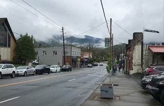 A small-town main street of low brick shopfronts and parked cars under a grey sky, with mountains behind.