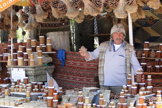 A moustached man in a flat cap and waistcoat stands behind tiered shelves of jars, a patterned kilim on the wall behind him.