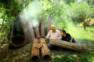 An older man and a girl sit on the ground working wicker into a long cylinder, finished cylinders standing and lying around them under fruit trees, smoke drifting through.