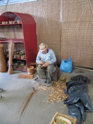 A man in a straw hat and grey jumper sits on a low wooden stand weaving a small basket, bundles of thin rods beside him and cut rod ends scattered on the concrete floor; woven screens and a red wicker shelf behind.