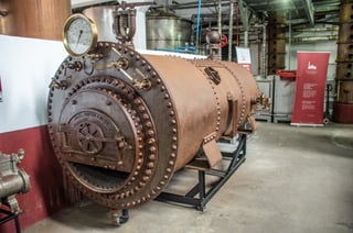 A large rusted riveted boiler on a black metal frame, a round brass pressure gauge on top and a cast door plate on its end; tall metal columns and pipes behind in a whitewashed hall.