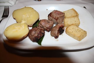 A white plate with four cubes of grilled beef on a leaf, two halves of a boiled potato and three golden fried cubes of maize porridge, on a white tablecloth beside a fork.