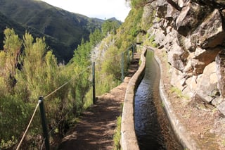 A narrow concrete water channel full of flowing water curves along a rocky hillside, a dirt path with green posts and a cable rail beside it, green valleys and mountains beyond.
