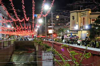 A night street scene: strings of red lights hang over a road beside a walled stream channel, crowds walk along it, and the lit facade of a building lettered Mercado dos Lavradores stands at the right with a hillside of lights behind.