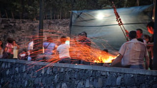 At dusk, orange sparks stream from an open fire behind a low stone wall, a skewer of meat raised above it on a pole, and people standing around in a crowd.
