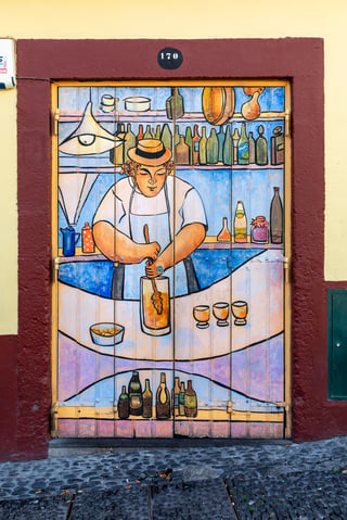 A pair of wooden doors in a dark red stone frame painted with a man in a straw hat and white apron stirring a yellow drink in a tall jug, shelves of bottles behind him and three small glasses on the counter; the number 170 above and cobbles below.