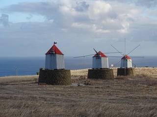 Three small wooden windmills with red pointed roofs standing on round dark stone bases on a dry grassy hilltop, two with wooden sail arms and wire rigging, the sea and a cloudy sky behind.