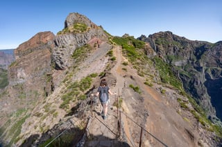 A walker with a backpack, seen from behind, goes down stone steps on a narrow ridge path with metal posts and cables, bare red and grey rock peaks rising ahead and deep valleys on either side under a clear sky.