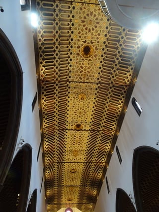 Looking straight up at a long wooden ceiling of gilded interlaced star and strap patterns, white walls and dark pointed arches on either side, spotlights at the top corners.