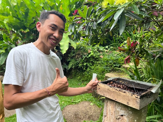 Wayan Indra stands beside an open beehive with a smile and a thumbs up.